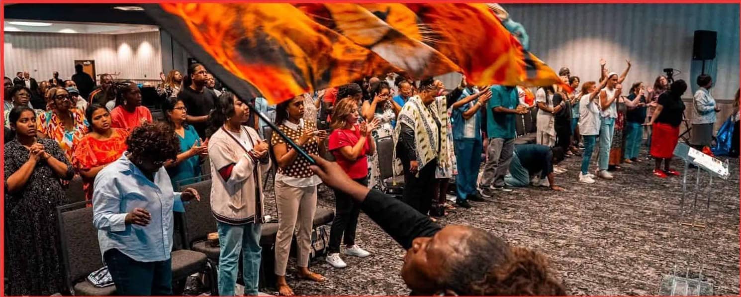Worshippers gathered in a revival service with a large red and orange flag waving in the foreground.